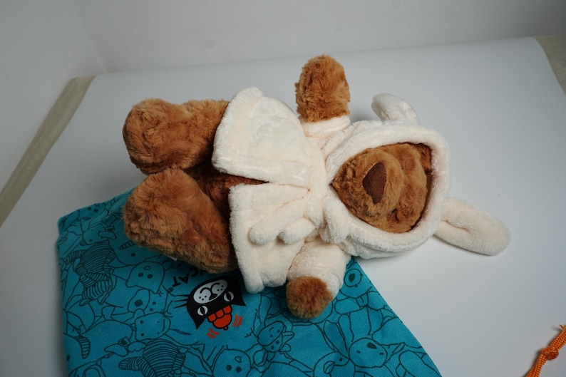 May include: A brown plush teddy bear wearing a cream-colored hooded robe with long ears attached to the hood. The bear is positioned on top of a teal fabric bag featuring a repeating pattern of black line-art cat faces and the word CAT.
