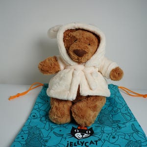 May include: A brown plush teddy bear wearing a cream-colored hooded bathrobe. The bear is positioned on a teal drawstring bag featuring a repeating black line-art pattern of various animals and the Jellycat logo printed in black at the bottom.