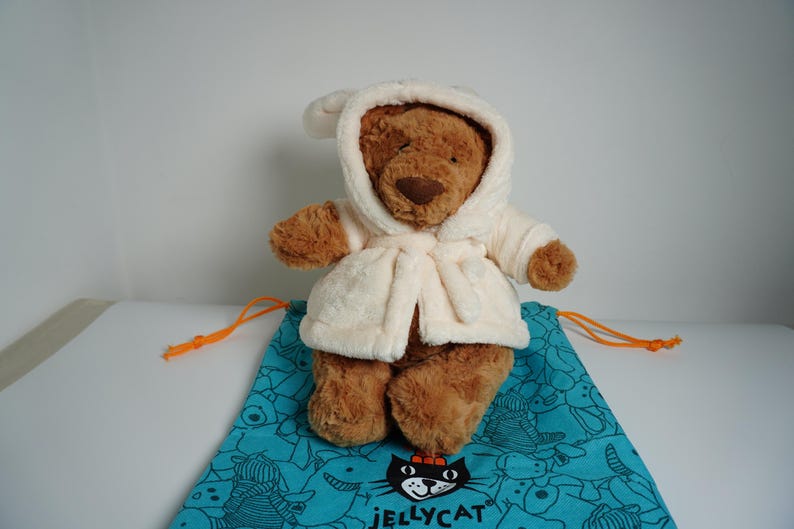 May include: A brown plush teddy bear wearing a cream-colored hooded bathrobe. The bear is positioned on a teal drawstring bag featuring a repeating black line-art pattern of various animals and the Jellycat logo printed in black at the bottom.
