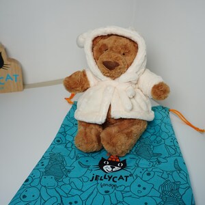 May include: A brown plush teddy bear wearing a cream-colored hooded robe, positioned on a teal drawstring bag featuring a black cat logo and the text JELLYCAT London. A wooden sign with the same cat logo and text is visible in the background.