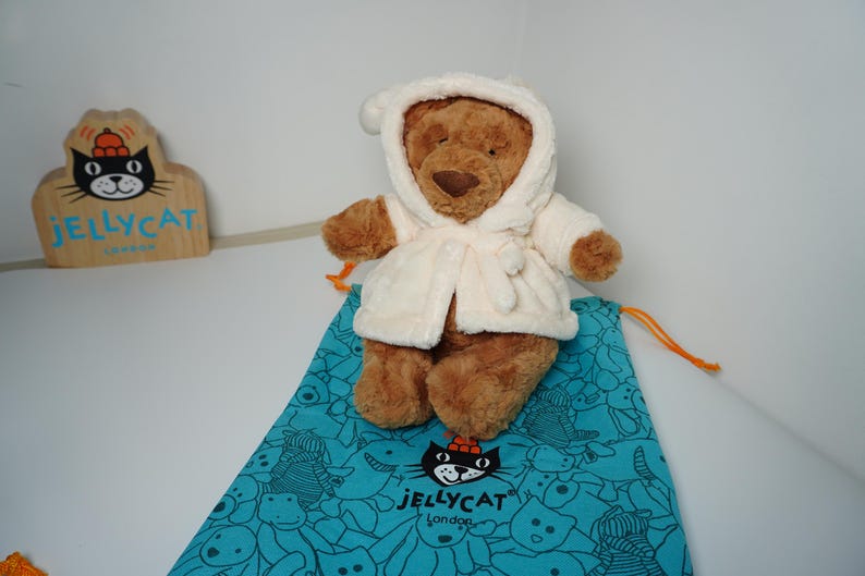May include: A brown plush teddy bear wearing a cream-colored hooded robe, positioned on a teal drawstring bag featuring a black cat logo and the text JELLYCAT London. A wooden sign with the same cat logo and text is visible in the background.