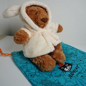 May include: A brown plush bear toy wearing a cream-colored hooded bathrobe. The bear is positioned on a turquoise drawstring bag featuring a repeating black line-art pattern of various animals and the text Jellycat London printed in black.