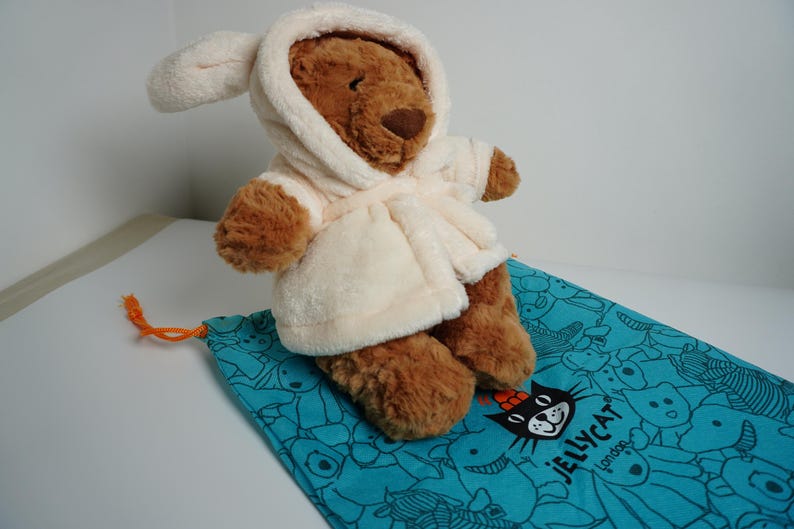 May include: A brown plush bear toy wearing a cream-colored hooded bathrobe. The bear is positioned on a turquoise drawstring bag featuring a repeating black line-art pattern of various animals and the text Jellycat London printed in black.