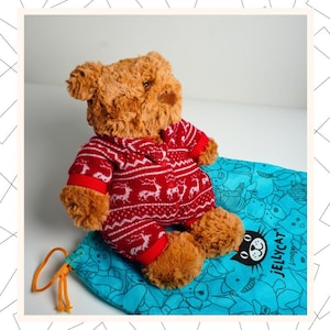 May include: A plush brown teddy bear wearing a red and white patterned onesie with a reindeer design. The bear sits on a blue drawstring bag with a cartoon print and the text "JELLYCAT London".