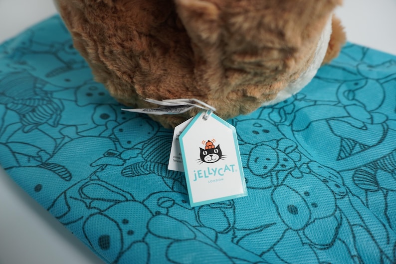 May include: A brown plush toy with a soft, textured surface resting on a bright turquoise fabric featuring a repeating black line-art pattern of various animals. A white tag with a black cat logo and the text JELLYCAT LONDON is attached.