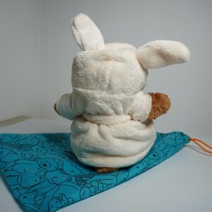 May include: A plush toy with long ears wearing a cream-colored hooded robe with a tie waist, positioned on a teal drawstring bag featuring a black line-art pattern of various small creatures and an orange cord.