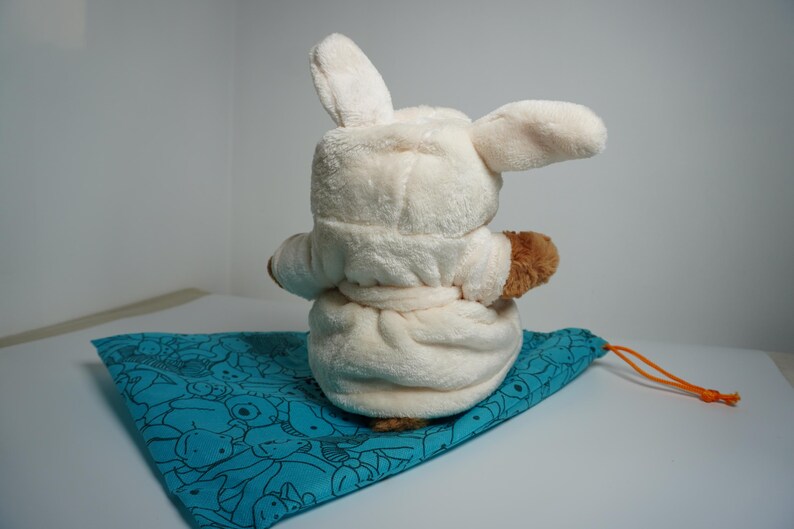 May include: A plush toy with long ears wearing a cream-colored hooded robe with a tie waist, positioned on a teal drawstring bag featuring a black line-art pattern of various small creatures and an orange cord.