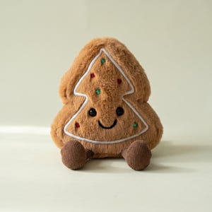 May include: A plush toy shaped like a brown gingerbread tree with a soft, fuzzy texture. It features a white embroidered outline, small red and green embroidered dots, black bead eyes, a stitched smile, and two small brown corduroy feet.