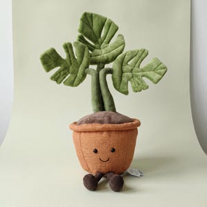 May include: A plush toy shaped like a potted monstera plant with three large green leaves. The orange fabric pot features a stitched smiling face with two black bead eyes and a curved mouth, along with two small dangling brown corduroy legs.