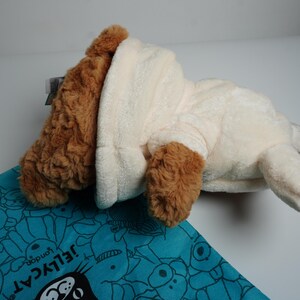 May include: A plush toy dog with brown fur on its head and ears, wearing a cream-colored hooded sweatshirt. The toy is positioned on a turquoise fabric bag featuring a repeating pattern of cartoon faces and the text Jellycat London.