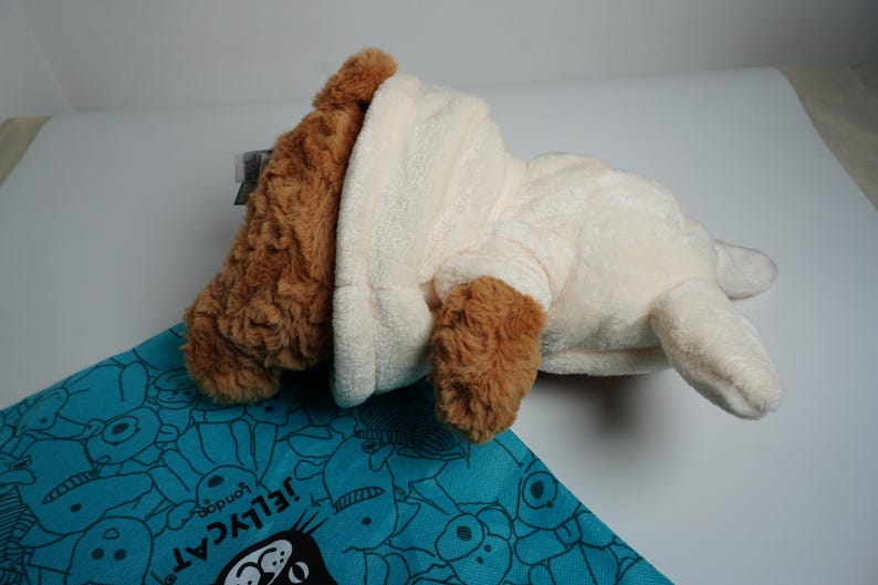 May include: A plush toy dog with brown fur on its head and ears, wearing a cream-colored hooded sweatshirt. The toy is positioned on a turquoise fabric bag featuring a repeating pattern of cartoon faces and the text Jellycat London.