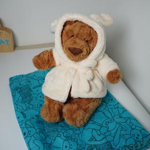 May include: A brown plush bear wearing a cream-colored hooded robe with ears on the hood. The bear is positioned on a teal drawstring bag featuring a repeating line-art pattern of various animals. A wooden Jellycat sign is visible in the background.