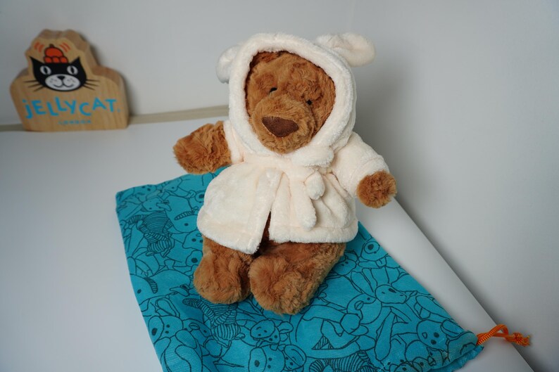 May include: A brown plush bear wearing a cream-colored hooded robe with ears on the hood. The bear is positioned on a teal drawstring bag featuring a repeating line-art pattern of various animals. A wooden Jellycat sign is visible in the background.