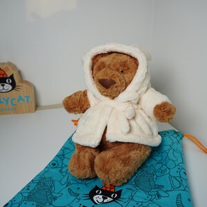 May include: A brown plush teddy bear wearing a cream-colored hooded robe with a tie closure. The bear is positioned on a teal drawstring bag featuring a black cat graphic. A wooden sign with the text JELLYCAT LONDON is visible in the background.