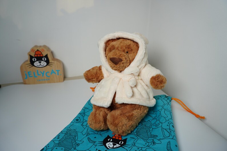 May include: A brown plush teddy bear wearing a cream-colored hooded robe with a tie closure. The bear is positioned on a teal drawstring bag featuring a black cat graphic. A wooden sign with the text JELLYCAT LONDON is visible in the background.