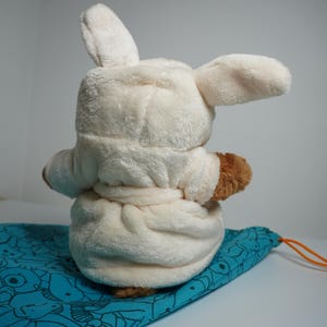 May include: A plush toy with long ears wearing a cream-colored hooded bathrobe with a tie waist. The toy has brown fur visible on its arms and is positioned on a teal drawstring bag featuring a repeating pattern of various animal line drawings.