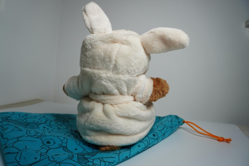 May include: A plush toy with long ears wearing a cream-colored hooded bathrobe with a tie waist. The toy has brown fur visible on its arms and is positioned on a teal drawstring bag featuring a repeating pattern of various animal line drawings.