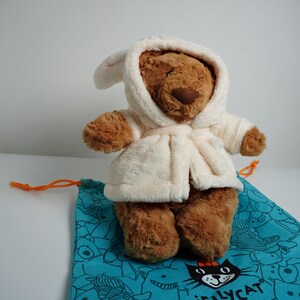 May include: A brown plush teddy bear wearing a cream-colored hooded bathrobe. The bear is positioned on a teal drawstring bag featuring a black cat graphic and the text Jellycat printed in black near the bottom edge of the fabric.