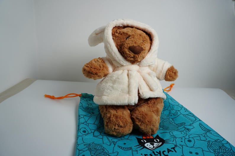 May include: A brown plush teddy bear wearing a cream-colored hooded bathrobe. The bear is positioned on a teal drawstring bag featuring a black cat graphic and the text Jellycat printed in black near the bottom edge of the fabric.
