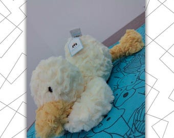 May include: A plush, cream-colored duck toy with a yellow beak and feet. The duck has a soft, fluffy texture and a small black eye. It is lying on a blue patterned surface. A tag is attached to the duck.