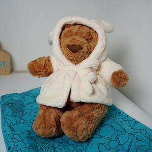 May include: A brown plush teddy bear wearing a cream-colored hooded robe with a tie closure. The bear has a dark brown nose and soft fur texture. In the background, a wooden sign features a black cat illustration and the text JELLYCAT LONDON.