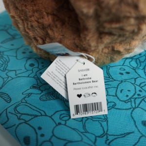 May include: A brown plush bear toy with a white tag reading I am Bathrobe Bartholomew Bear Please look after me with a heart, bear, and person icon, resting on a bright turquoise fabric featuring a repeating black line drawing pattern of bears.