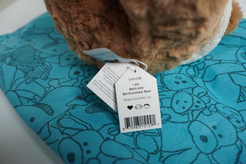 May include: A brown plush bear toy with a white tag reading I am Bathrobe Bartholomew Bear Please look after me with a heart, bear, and person icon, resting on a bright turquoise fabric featuring a repeating black line drawing pattern of bears.