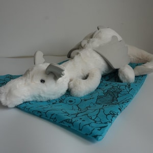May include: A white plush dragon toy with smooth gray horns and wings, resting on a bright teal fabric square featuring a black line-art pattern of various small creatures and shapes, including a prominent striped character design.
