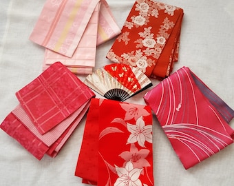 Puede incluir: Una colección de cinco fajas de tela rectangulares plegadas en tonos rosa y rojo, con varios patrones que incluyen estampados florales, rayas y cuadrículas geométricas, dispuestas alrededor de un pequeño abanico abierto con detalles en oro y rojo.