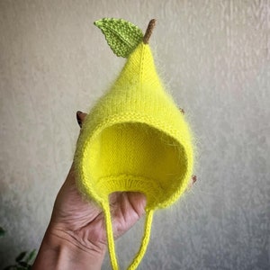 Puede incluir: Un gorro de bebé de punto amarillo con forma de pera y una hoja verde en la parte superior.