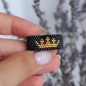 Puede incluir: Un anillo de cuentas negras con un diseño de corona dorada. El anillo está en un dedo.