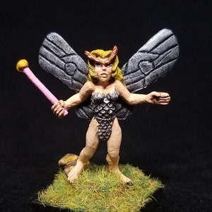 May include: A hand-painted miniature figurine of a fairy with silver wings, blonde hair, and a pink wand. The fairy is wearing a silver and black scale-like outfit and is standing on a green grassy base.