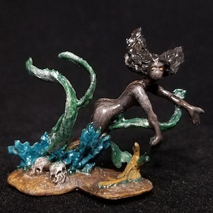 Peut inclure: Une figurine peinte en gris foncé et vert d'une femme aux cheveux longs, debout dans une flaque d'eau avec des crânes. La figurine est en résine et mesure environ 10 cm de haut.