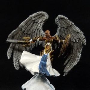 May include: A painted miniature figurine of an angel with silver wings, a white and blue robe, and a golden sword. The angel is standing on a white cloud base.