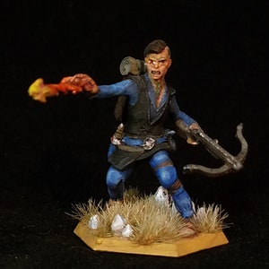 Op de afbeelding: Een geschilderd miniatuurfiguur van een persoon in blauwe kleding die een kruisboog vasthoudt. Het figuur staat op een bruine basis met een paar witte stenen en gras.