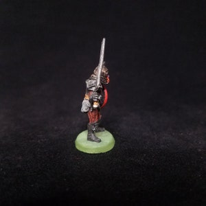 Pu&ograve; includere: Una figura in miniatura dipinta di un guerriero Orco che tiene una spada. L'Orco indossa un'armatura nera e abiti rossi. La figura &egrave; in piedi su una base verde.