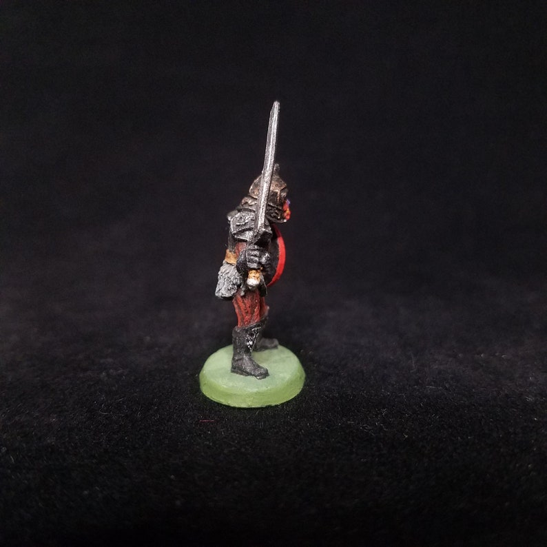 Pu&ograve; includere: Una figura in miniatura dipinta di un guerriero Orco che tiene una spada. L'Orco indossa un'armatura nera e abiti rossi. La figura &egrave; in piedi su una base verde.