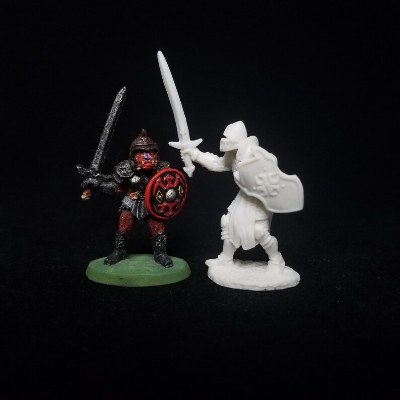 Pu&ograve; includere: Una figura in miniatura dipinta di rosso e nero di un guerriero che tiene una spada e uno scudo, di fronte a una figura in miniatura bianca non dipinta di un guerriero che tiene una spada e uno scudo.