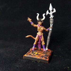 Puede incluir: Figura de fantasía en miniatura de una demonio femenina con cuernos, cola y atuendo morado, sosteniendo un tridente plateado. La figura se alza sobre una base cuadrada con textura de lava, sobre un fondo oscuro.