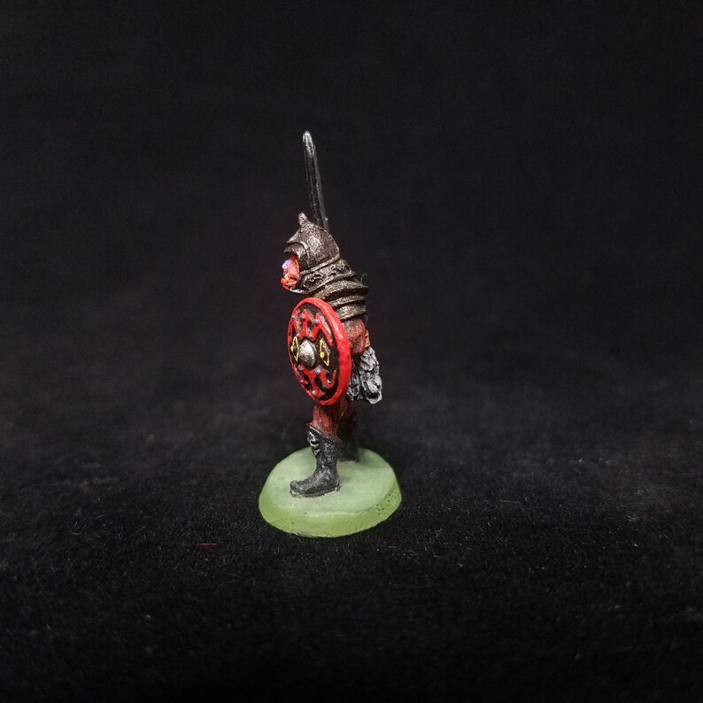 Pu&ograve; includere: Una figura in miniatura dipinta di un guerriero che indossa un'armatura nera e tiene uno scudo rosso con accenti dorati. La figura &egrave; in piedi su una base verde.