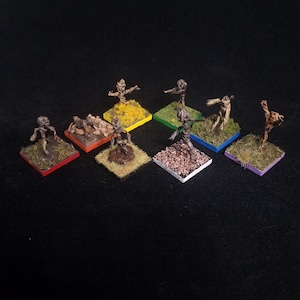 May include: Eight miniature figures made of twigs and branches, each standing on a small square base with a different colored background. The figures are arranged in a circle on a black surface.