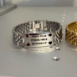 Puede incluir: Pulsera de metal plateado con la inscripci&oacute;n "PIKES PEAK POKER TOUR RUNNER UP" y s&iacute;mbolos de cartas. La pulsera tiene un dise&ntilde;o de eslabones y un cierre. Una pulsera dorada es parcialmente visible.