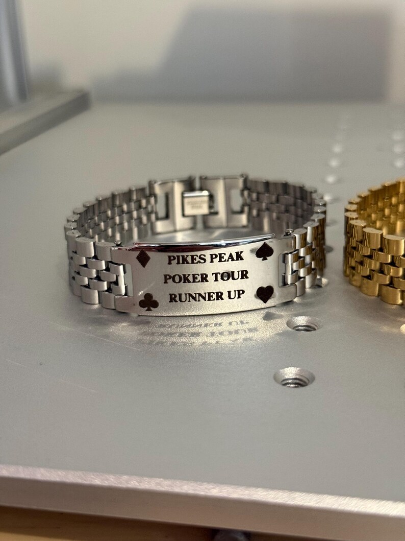 Puede incluir: Pulsera de metal plateado con la inscripci&oacute;n "PIKES PEAK POKER TOUR RUNNER UP" y s&iacute;mbolos de cartas. La pulsera tiene un dise&ntilde;o de eslabones y un cierre. Una pulsera dorada es parcialmente visible.