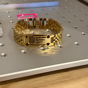 Puede incluir: Una pulsera dorada con una placa rectangular grabada con el texto "Big Brothers Big Sisters of Snohomish County" y "POKER CHAMPION 2025". La pulsera presenta un dise&ntilde;o de cadena de eslabones y est&aacute; sobre una superficie plateada.