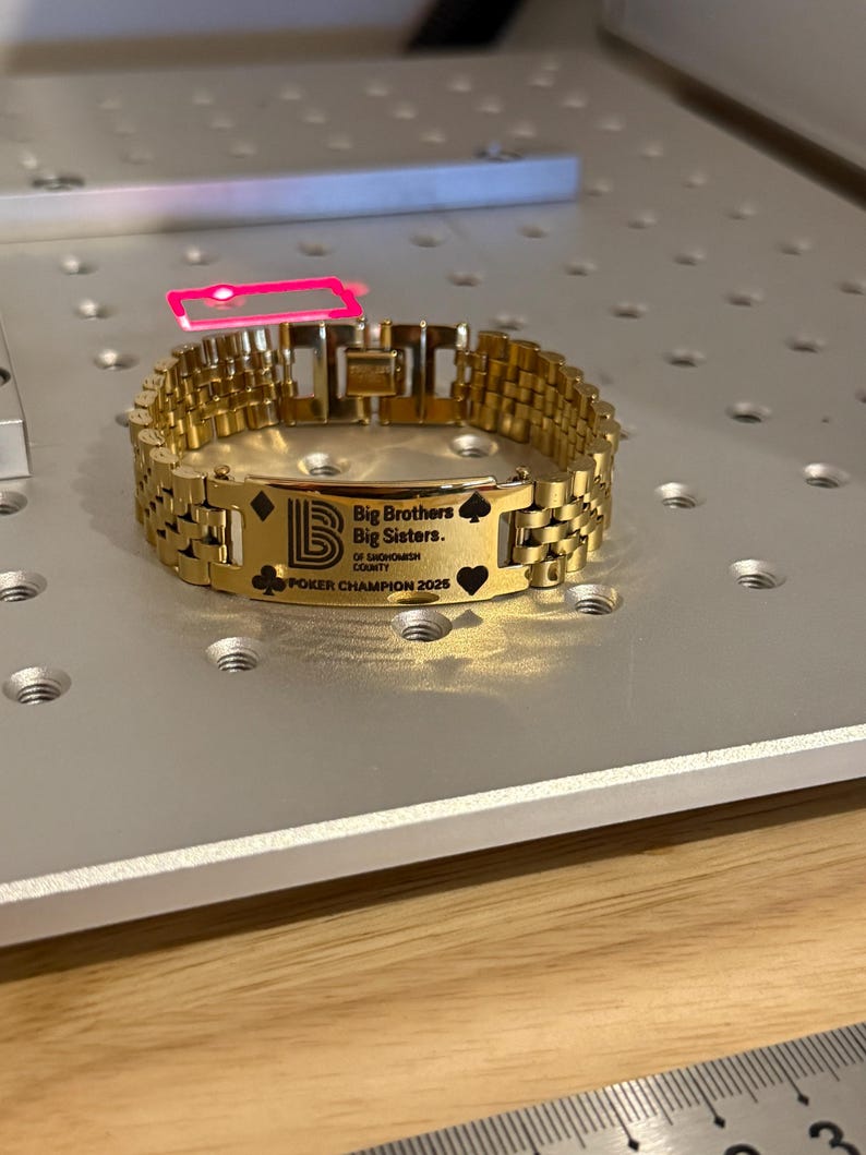 Puede incluir: Una pulsera dorada con una placa rectangular grabada con el texto "Big Brothers Big Sisters of Snohomish County" y "POKER CHAMPION 2025". La pulsera presenta un dise&ntilde;o de cadena de eslabones y est&aacute; sobre una superficie plateada.