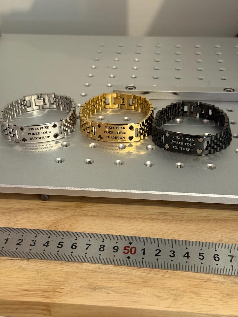 Puede incluir: Tres pulseras met&aacute;licas en plata, oro y negro, cada una grabada con "PIKES PEAK POKER TOUR" y una clasificaci&oacute;n: Runner Up, Champion y Top Three. Las pulseras se exhiben sobre una superficie gris claro con un fondo de madera.