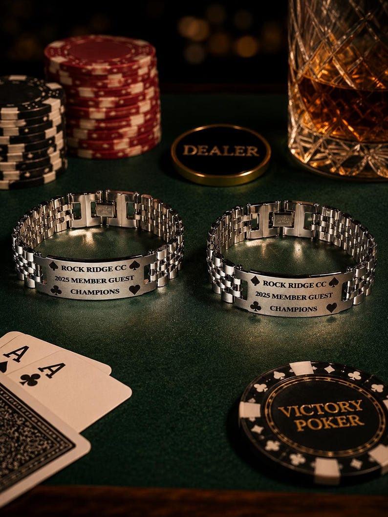 Puede incluir: Dos pulseras plateadas con la inscripci&oacute;n "ROCK RIDGE CC 2025 MEMBER GUEST CHAMPIONS" se exhiben sobre una superficie verde. Fichas de p&oacute;quer, naipes y un vaso de l&iacute;quido &aacute;mbar sugieren un tema de p&oacute;quer.