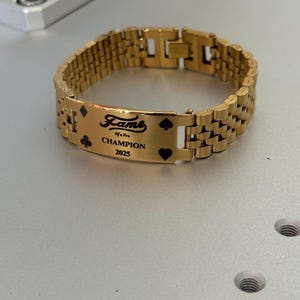 Puede incluir: Pulsera dorada con una placa rectangular con la palabra "Fame" y el texto "Champion 2025". La placa tambi&eacute;n incluye s&iacute;mbolos de naipes. La pulsera tiene un dise&ntilde;o de eslabones.