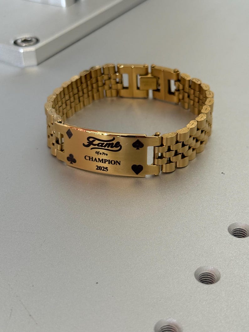 Puede incluir: Pulsera dorada con una placa rectangular con la palabra "Fame" y el texto "Champion 2025". La placa tambi&eacute;n incluye s&iacute;mbolos de naipes. La pulsera tiene un dise&ntilde;o de eslabones.