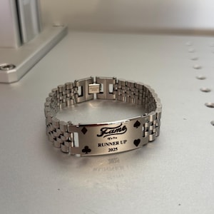 Puede incluir: Una pulsera de metal plateado con una placa rectangular grabada con la palabra "Fame", el texto "of a Pro", "Runner Up" y el a&ntilde;o "2025". La placa tambi&eacute;n presenta s&iacute;mbolos de palos de cartas. La pulsera tiene un dise&ntilde;o de eslabones.